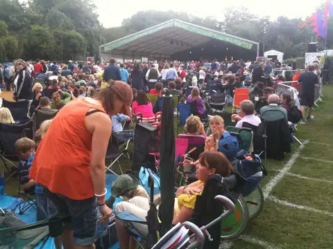 Audience in folding chairs on grass in front of a large stage tent among trees