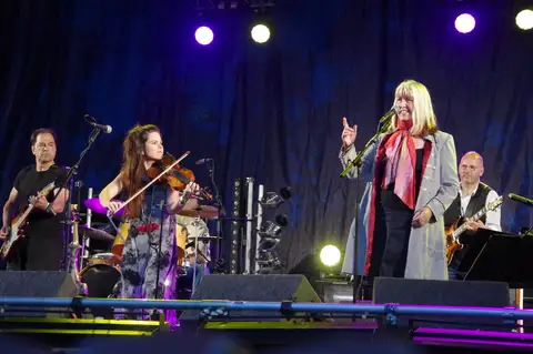 A folk-rock band performing on a stage lit in blue and purple, with a fiddler and a singer at the front