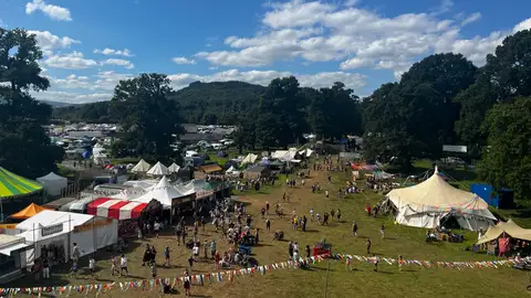 A festival site of white tents and marquees in a green valley ringed by wooded hills