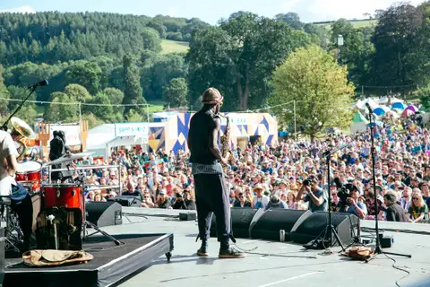 Crowd on a sunny hillside facing a festival main stage with a percussionist in the foreground