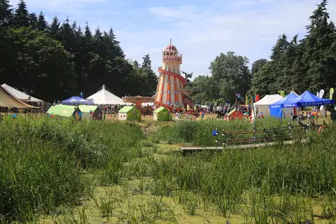 A lake fringed with tall reeds, with festival tents and a striped helter-skelter among the trees beyond